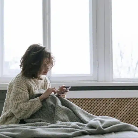 A woman sitting on a bed by a large window, wrapped in a blanket while using her phone.