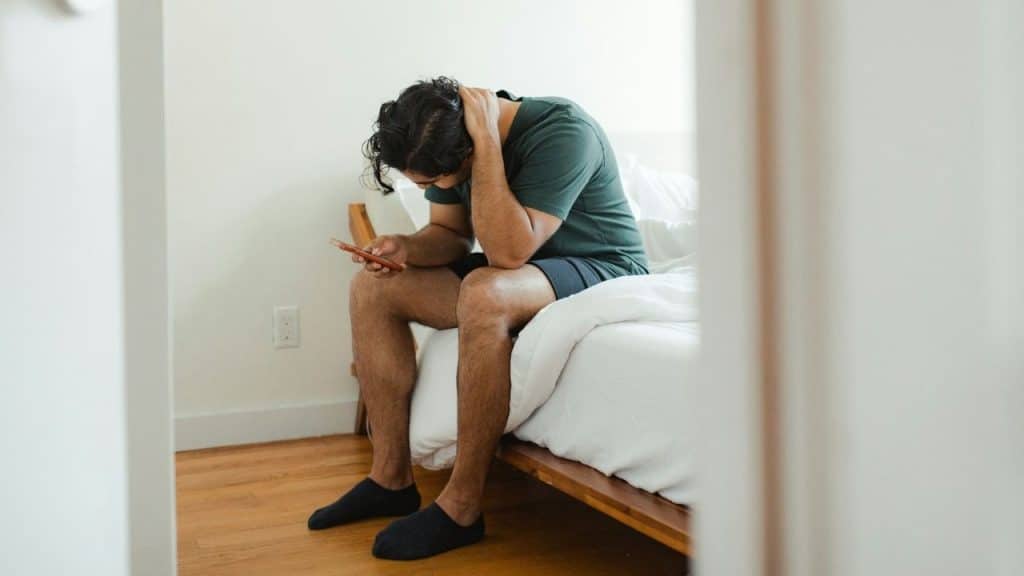 A man sits on the edge of a bed looking down at a mobile phone in his hand.