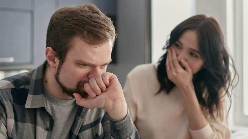 A bearded man looks down while a woman behind him covers her mouth with her hand.