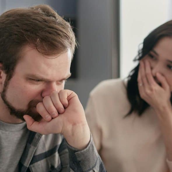 A bearded man looks down while a woman behind him covers her mouth with her hand.
