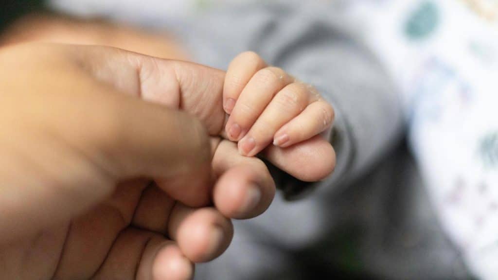 A baby holding an adult’s finger in a close-up shot.