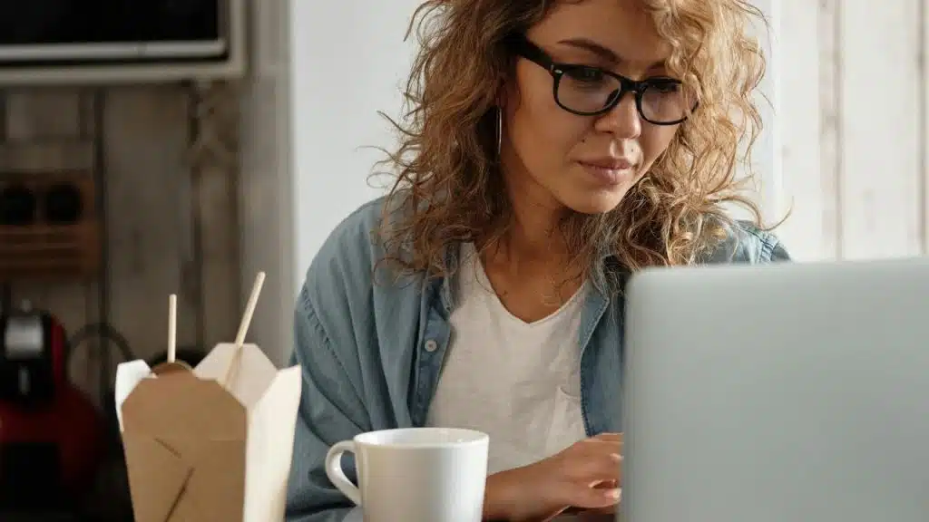 A woman wearing glasses working on a laptop with a coffee cup nearby.