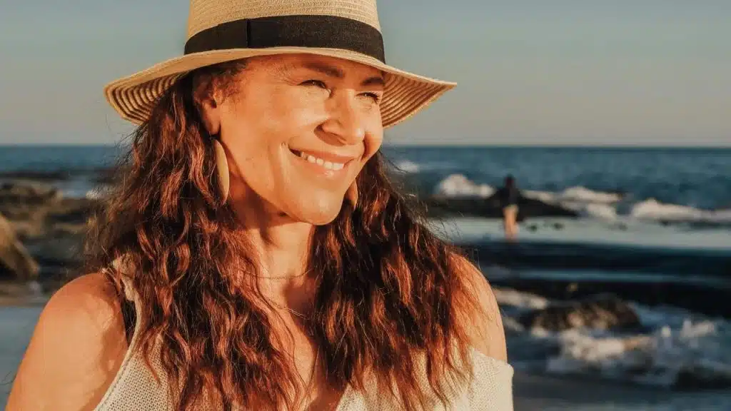 A smiling woman in a sun hat standing by the ocean at sunset.