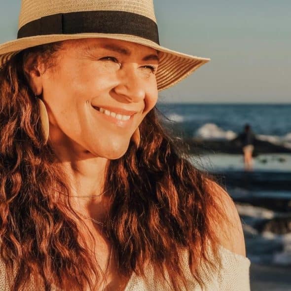 A smiling woman in a sun hat standing by the ocean at sunset.