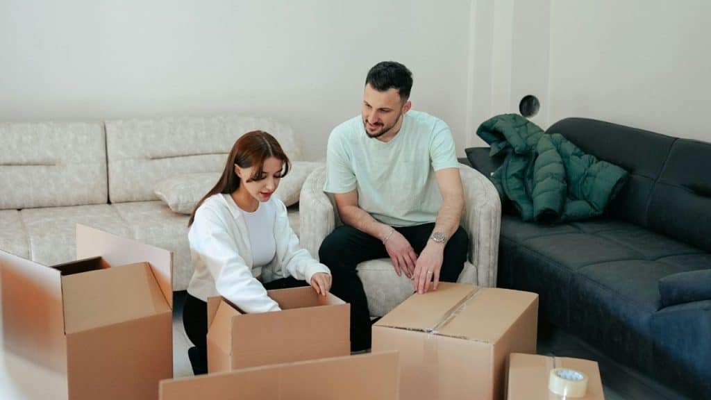 A man and woman sit among several open cardboard boxes in a living room setting.