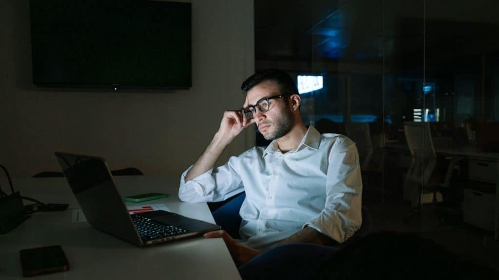 A man wearing glasses sits in a dark office looking at a glowing laptop screen.