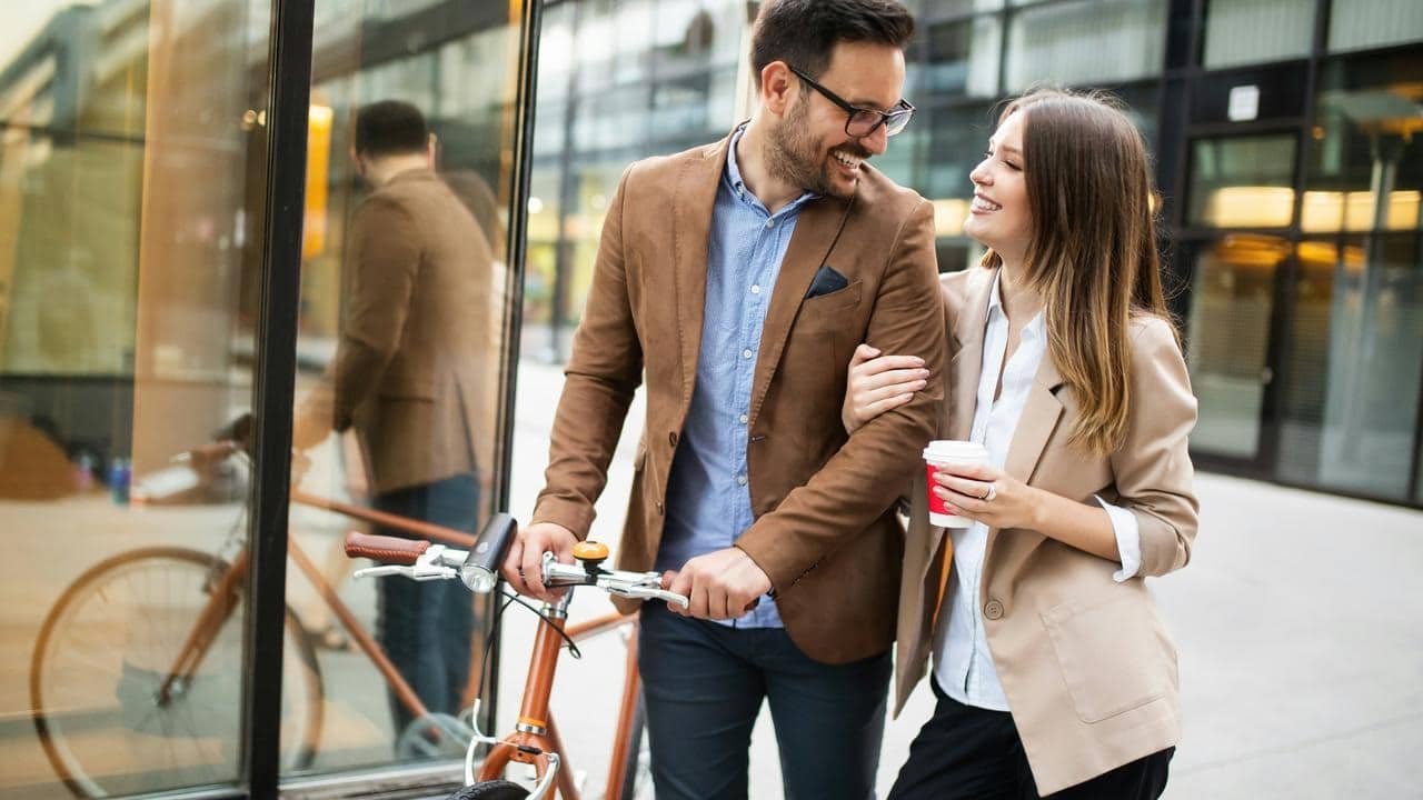 A smiling couple walking together with a bicycle and coffee.