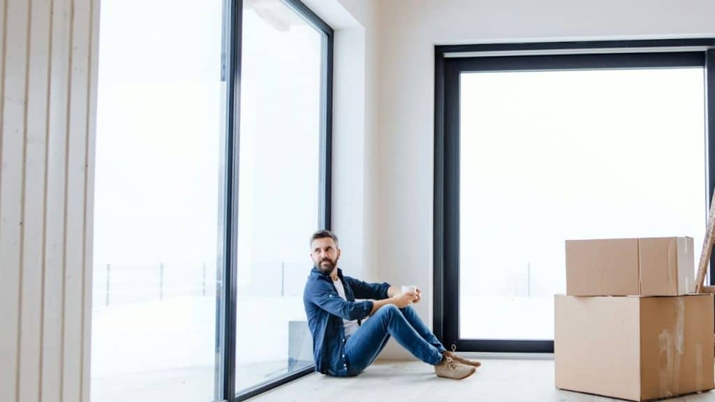 A bearded man sits on the floor near large windows and cardboard boxes holding a mug.