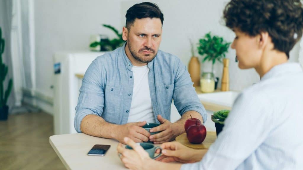 A man and woman sit at a table with mugs and apples, looking at each other.