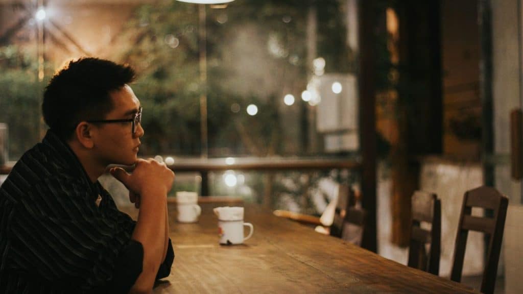 A man wearing glasses sits alone at a long wooden table in a dimly lit room.