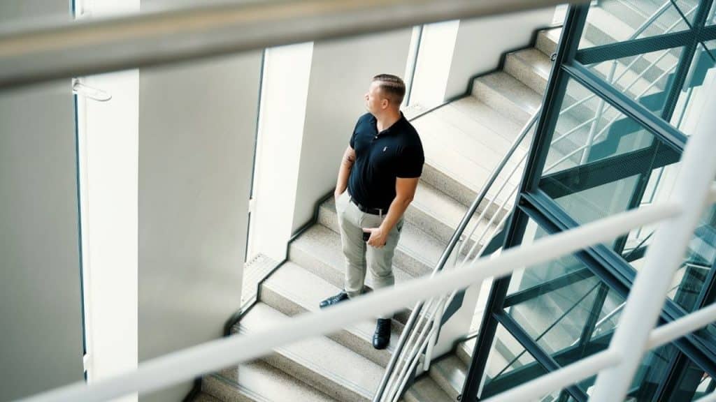 A man stands on a modern staircase looking out of a tall window while holding a phone.
