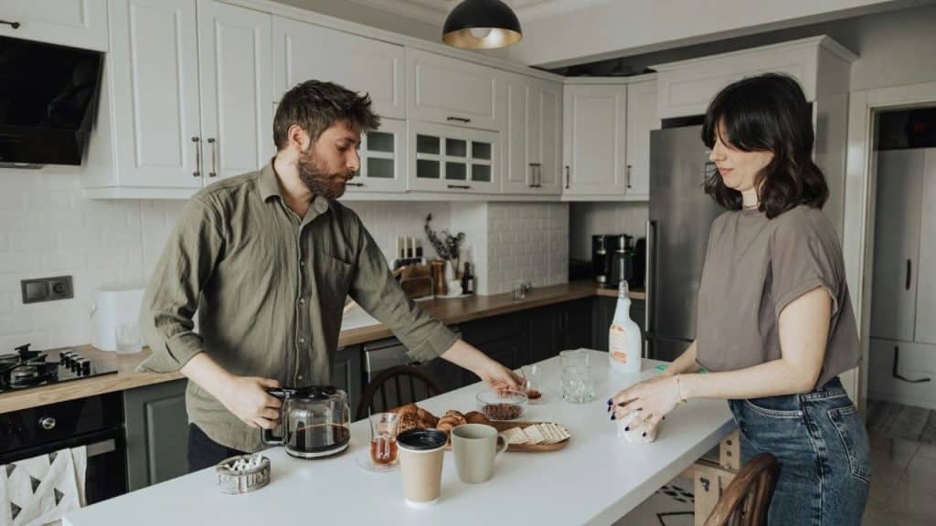 A man and woman stand in a kitchen preparing coffee and snacks on a white island.