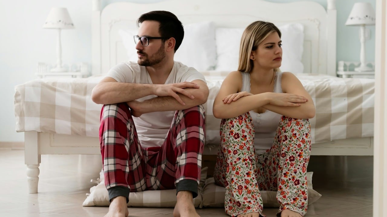 A couple in pajamas sitting on the floor are looking away from each other.
