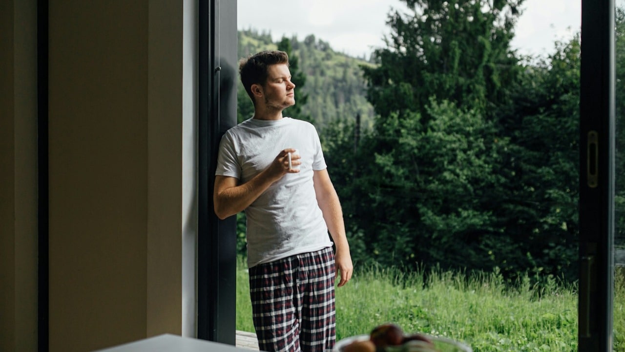 A man in a white T-shirt and plaid pajama pants stands in an open doorway holding a mug, looking outside toward a green, tree-filled landscape.