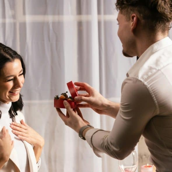 A man wearing a white shirt is proposing to his girlfriend with a box of ring on a romantic date.