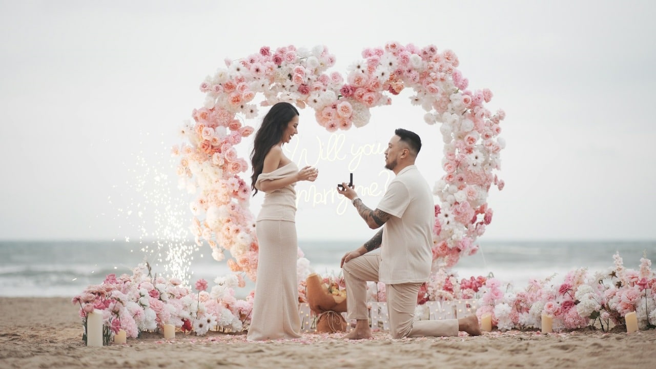 A man proposes to a woman on a beach with a heart arch made up of flowers.