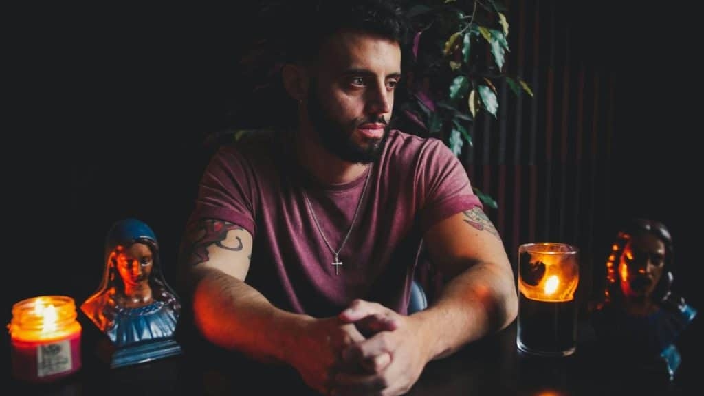 A bearded man sits at a dark table with folded hands, candles, and religious figurines.