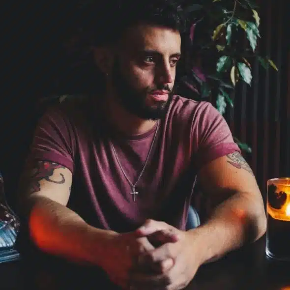 A bearded man sits at a dark table with folded hands, candles, and religious figurines.