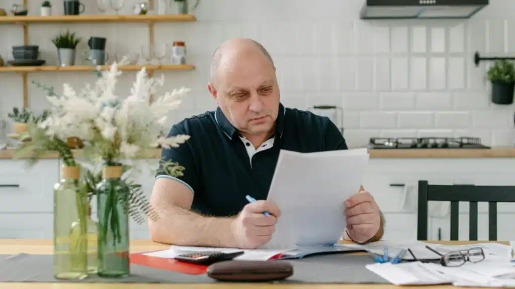 A man sits at a kitchen table holding a paper and pen near a calculator.