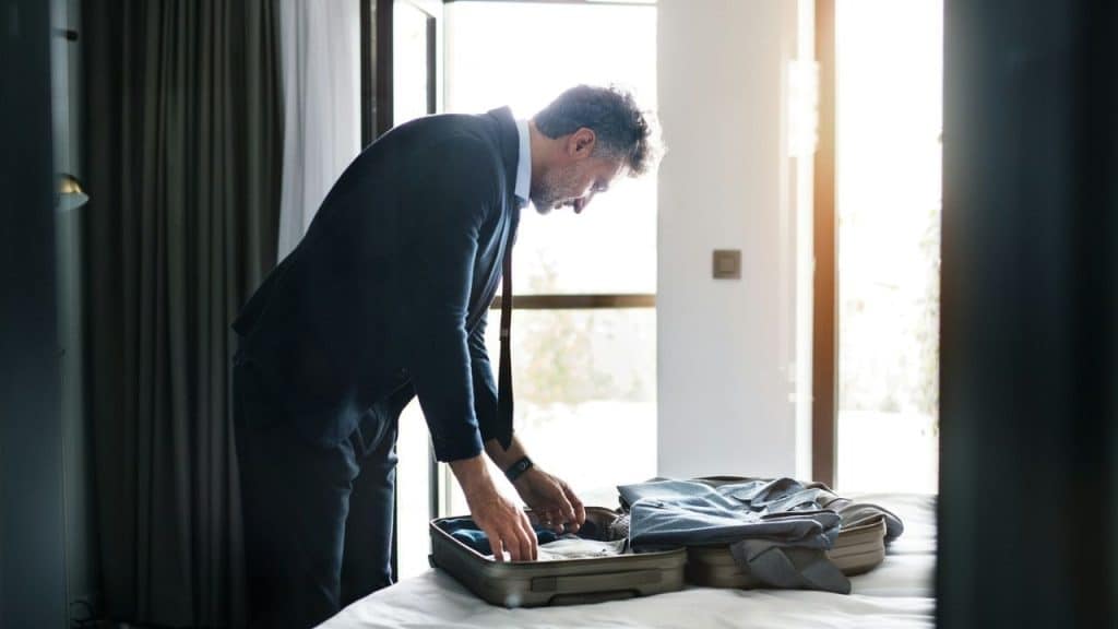 A man in a suit bends over a bed to pack clothes into a suitcase.