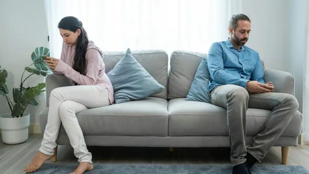 A woman and man sit far apart on a couch, looking at their individual phones.