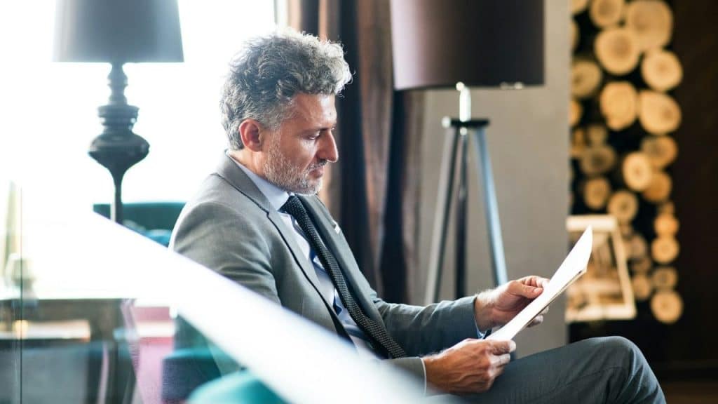 A man in a gray suit sits indoors and reads a white piece of paper.
