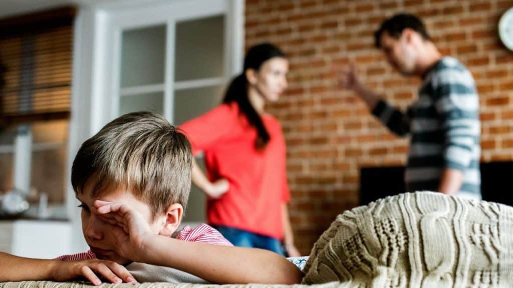 A child rubs his eye in the foreground while two adults stand blurred behind him.
