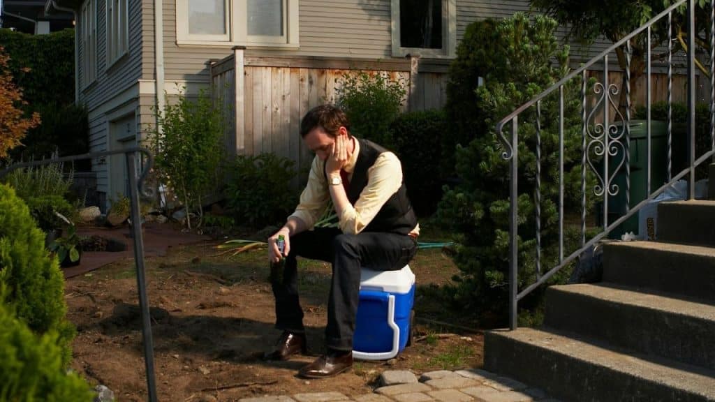A man sits outside on a blue cooler with his head resting in his hand.