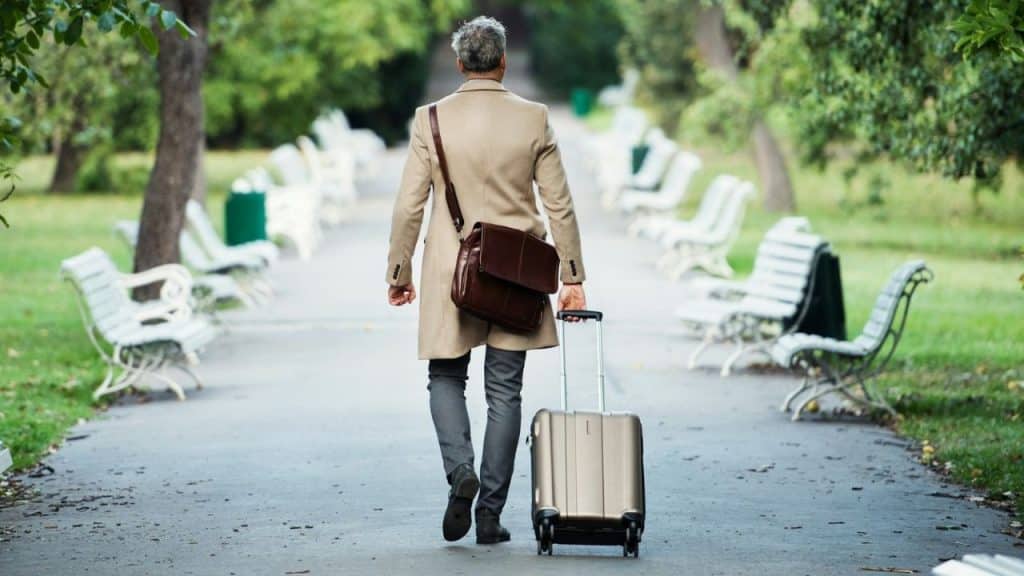 A man walks down a park path away from the camera, pulling a silver suitcase.