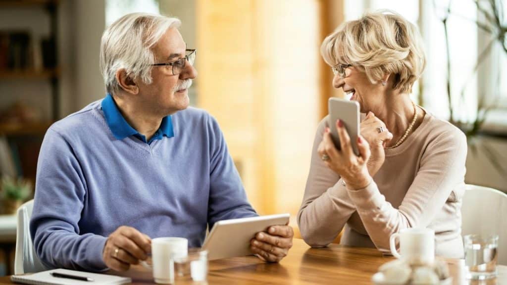 An older man and woman sit at a wooden table using a tablet and smartphone.