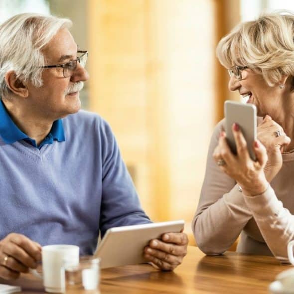 An older man and woman sit at a wooden table using a tablet and smartphone.