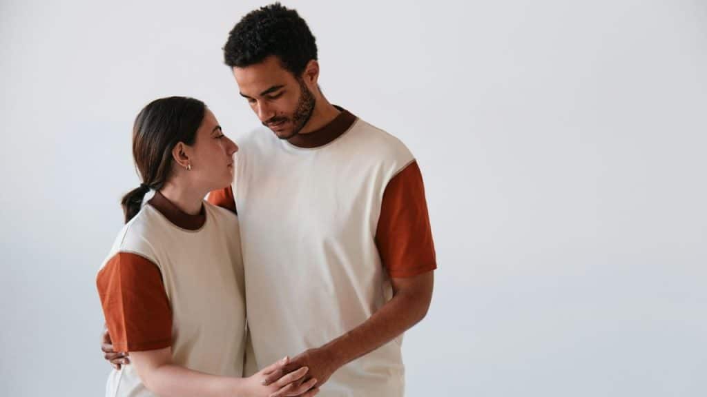 A man and woman stand together against a white background holding hands and looking down.