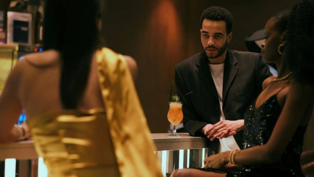 A man at a bar looks toward two women sitting nearby in a dimly lit setting.