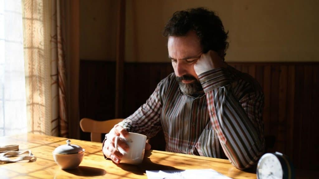 A bearded man sits at a wooden table holding a mug with his head downcast.