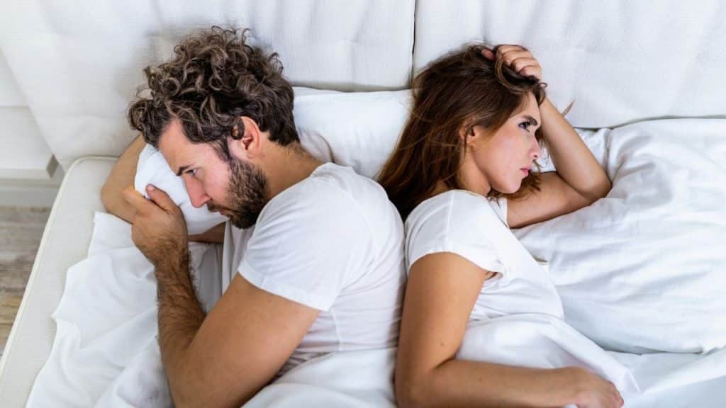 A man and woman lie in bed facing away from each other on white pillows.