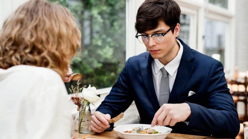 A man in a suit and glasses eats dinner while a woman sits across from him.