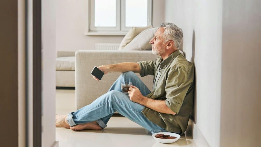 A man sits on the floor leaning against a wall while holding a phone and glass.