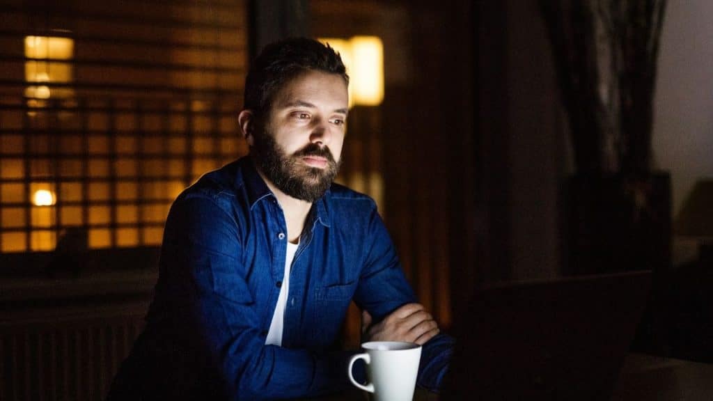 A bearded man in a blue shirt sits at a dark table with a white mug.