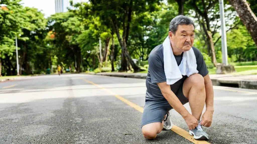 A man in athletic wear kneels on an outdoor path to tie his running shoe.