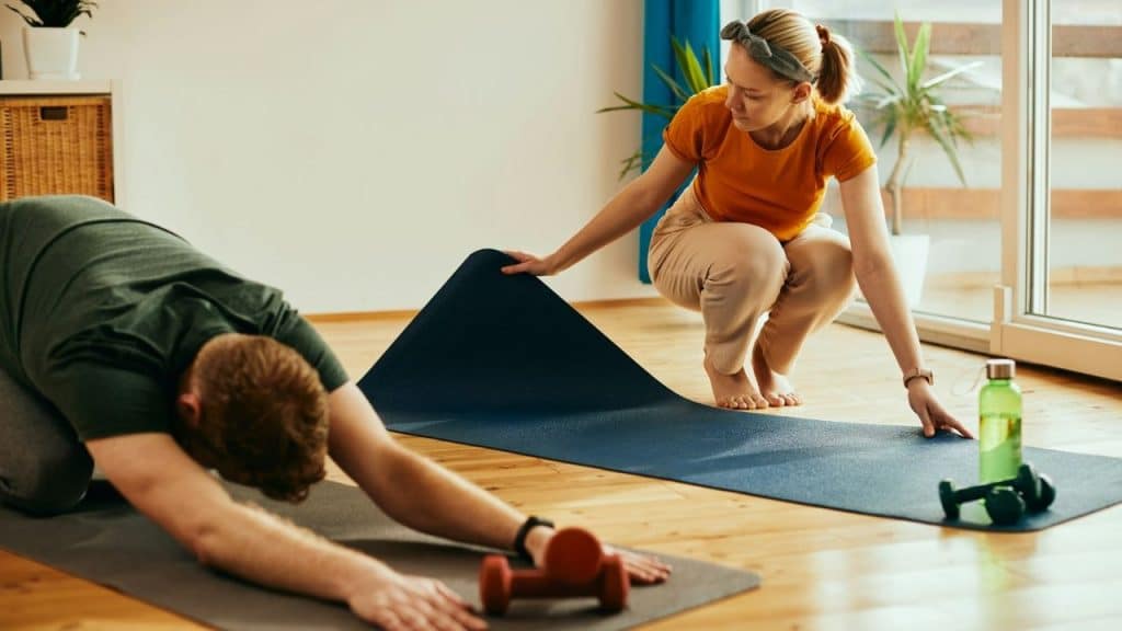 A woman in an orange shirt unrolls a yoga mat while a man stretches nearby.