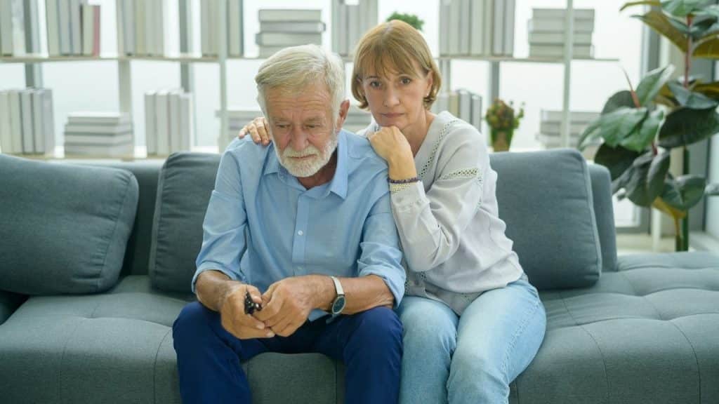 An older woman rests her hand on the shoulder of a man looking down on a sofa.