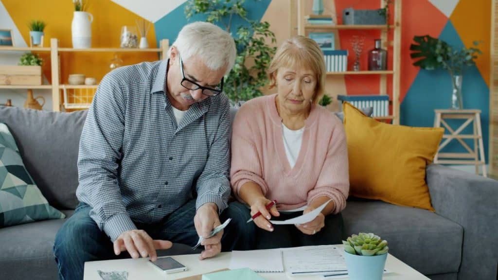 An older couple sits on a sofa looking at papers and a smartphone while holding money.