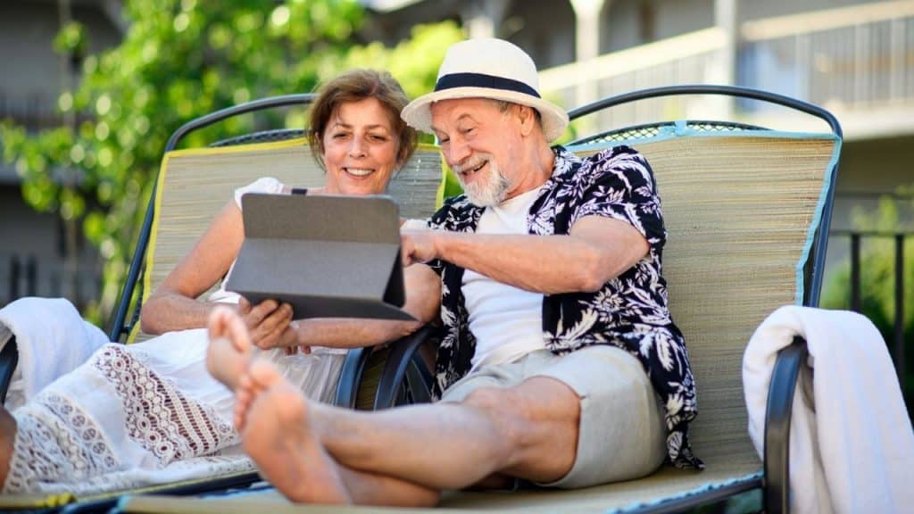 A smiling older couple sits in lounge chairs outdoors looking at a tablet together.