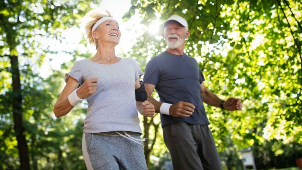 An older man and woman in athletic wear run together through a sunlit park.
