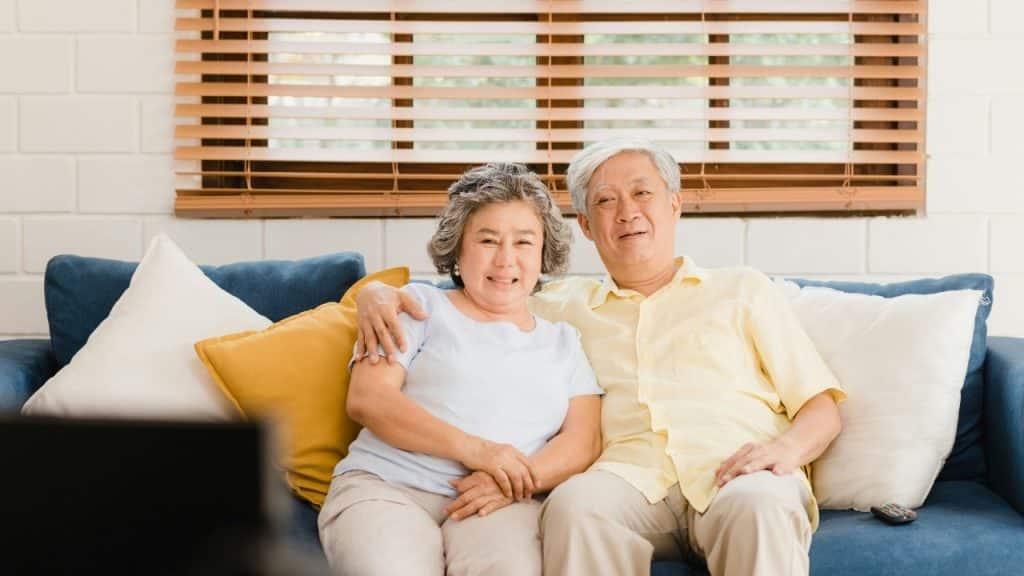 An older man and woman sitting together on a blue couch in front of a window.