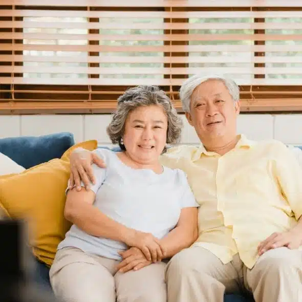 An older man and woman sitting together on a blue couch in front of a window.