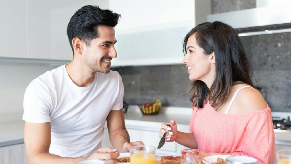 A man and woman smiling at each other while sitting at a kitchen table with breakfast.