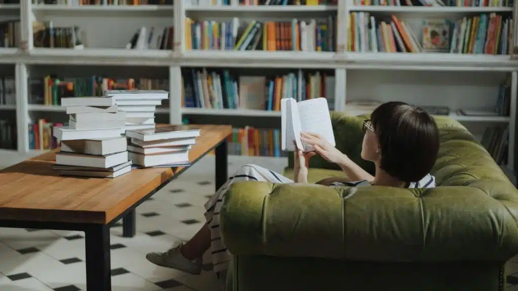 A woman sitting in a green leather armchair reading a book in a large library.