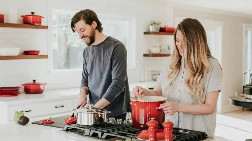 A man and woman smiling while cooking together at a stove in a bright kitchen.