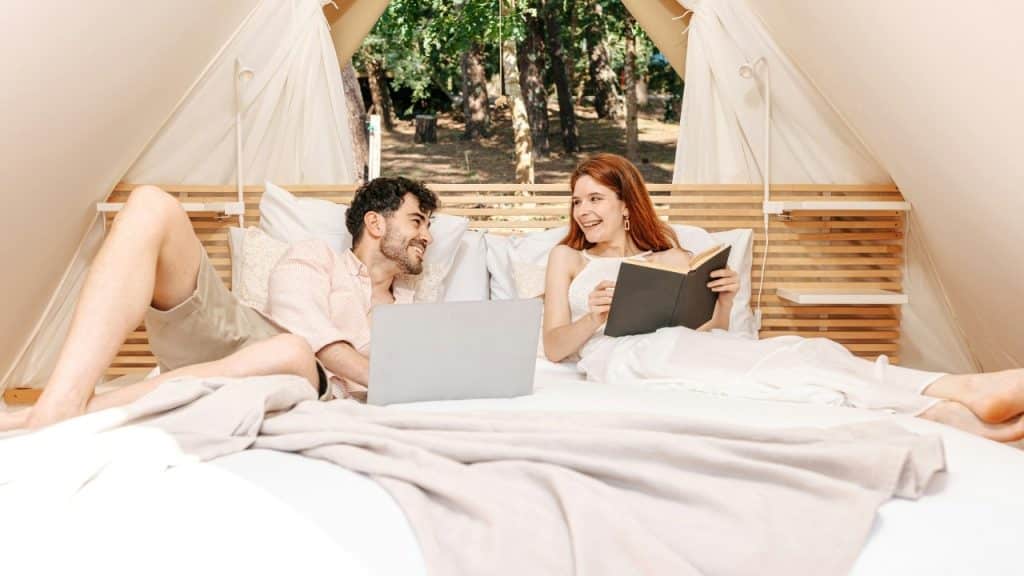 A man and woman sitting on a bed inside a tent, using a laptop and book.